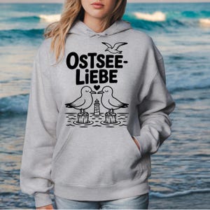 Könnte beinhalten: Ein hellgrauer Hoodie mit dem Text "OSTSEE-LIEBE" in fetten schwarzen Buchstaben. Das Design zeigt zwei Möwen, einen Leuchtturm und eine fliegende Möwe. Der Hoodie hat eine Vordertasche und eine Kapuze.