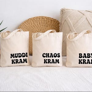 Könnte beinhalten: Drei cremefarbene Canvas-Tragetaschen mit schwarzem Text: "MUDDI KRAM", "CHAOS KRAM" und "BABY KRAM". Die Taschen haben Henkel. Im Hintergrund sind eine Topfpflanze, eine runde Webmatte und ein dekoratives Kissen zu sehen.