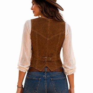 May include: A brown suede vest with a buckle belt, worn with a brown cowboy hat and blue jeans. The vest features a V-shaped design on the back. The outfit is completed with a white blouse with lace trim.