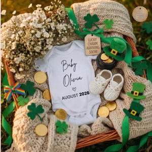 以下が含まれることがあります： 「Baby Olivia」と「August 2026」の日付が入った白いベビーボディスーツは、聖パトリックの日のテーマの装飾に囲まれています。画像には、金のポット、緑の帽子、シャムロック、ベビーシューズが含まれています。