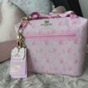 K&ouml;nnte beinhalten: Eine hellrosa Handtasche mit einem sich wiederholenden Muster aus kleinen wei&szlig;en Symbolen und dem auf der Vorderseite aufgedruckten Text "Juicy Couture". Die Tasche verf&uuml;gt &uuml;ber eine goldfarbene Metallkettenbefestigung mit passendem Anh&auml;nger.