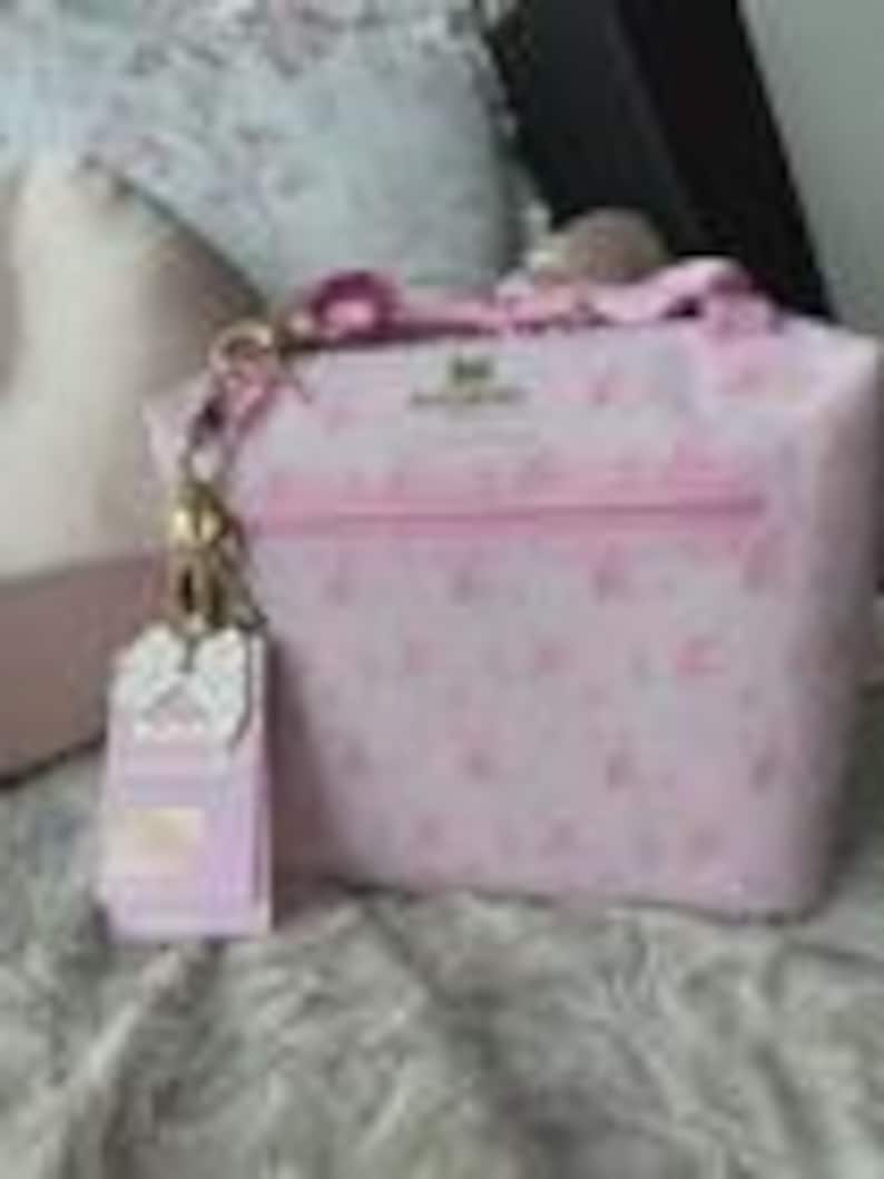 K&ouml;nnte beinhalten: Eine hellrosa Handtasche mit einem sich wiederholenden Muster aus kleinen wei&szlig;en Symbolen und dem auf der Vorderseite aufgedruckten Text "Juicy Couture". Die Tasche verf&uuml;gt &uuml;ber eine goldfarbene Metallkettenbefestigung mit passendem Anh&auml;nger.