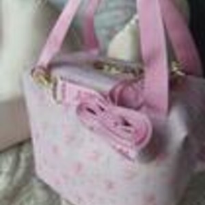 K&ouml;nnte beinhalten: Eine hellrosa, quadratische Tasche mit einem wiederkehrenden Muster aus kleinen rosa Blumenmotiven. Die Tasche verf&uuml;gt &uuml;ber zwei rosa Tragegriffe aus Stoff und einen abnehmbaren rosa Schulterriemen mit goldfarbenen Metallverschl&uuml;ssen.