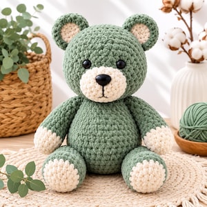 Op de afbeelding: Een gehaakte teddybeer gemaakt van saliegroen garen met crèmekleurige accenten op de oren, snuit en poten. Het knuffeldier heeft ronde zwarte plastic ogen en een kleine zwarte plastic neus, met een getextureerde, zacht ogende garenstructuur.