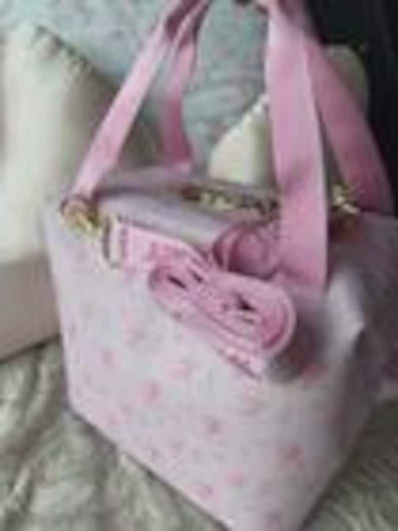 K&ouml;nnte beinhalten: Eine hellrosa, quadratische Tasche mit einem wiederkehrenden Muster aus kleinen rosa Blumenmotiven. Die Tasche verf&uuml;gt &uuml;ber zwei rosa Tragegriffe aus Stoff und einen abnehmbaren rosa Schulterriemen mit goldfarbenen Metallverschl&uuml;ssen.