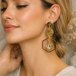 May include: Gold-toned earrings with a geometric design. The earrings feature a black and gold textured stud, a textured gold disc, a black hexagon, and a heart-shaped pendant with a clear stone. The earrings are a statement piece.