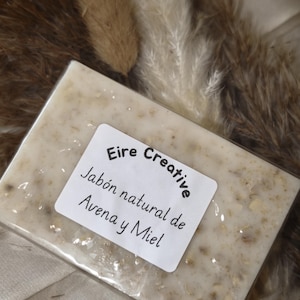 Puede incluir: Una pastilla de jabón rectangular de avena y miel con una superficie texturizada que contiene pequeños copos de avena. Una etiqueta blanca está centrada en la pastilla con el texto negro Eire Creative, Jabón natural de Avena y Miel.
