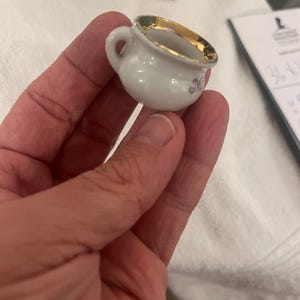 May include: A small, white porcelain chamber pot with a gold rim and handle. The miniature pot has a delicate design and is likely a collectible or decorative item. The pot has the letter "M" on the side.