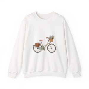 May include: A white crewneck sweatshirt featuring a centered graphic illustration of a light green bicycle. The bicycle has a brown leather seat, a brown rear pannier bag, and a front wicker basket filled with a bouquet of sunflowers and lavender.