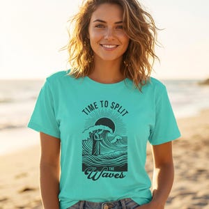 Puede incluir: Una camiseta turquesa con el texto "TIME TO SPLIT THE WAVES" en negro. El gráfico presenta un sol, olas y una figura. La camiseta es de manga corta y está hecha de un material suave.