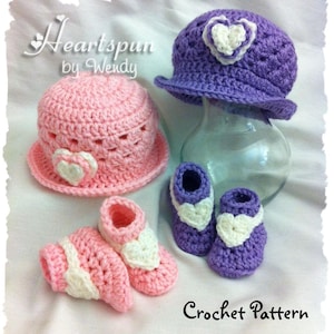 Puede incluir: Un conjunto de gorro y patucos de ganchillo para bebé en rosa y morado con detalles de corazón blanco. El conjunto incluye un gorro con ala y patucos con un diseño de corazón en la parte delantera. El patrón de ganchillo está disponible para su compra.