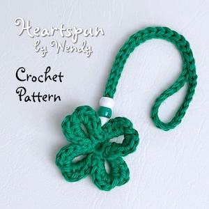 Pode incluir: Encanto de flor de crochê verde com uma conta branca e verde presa a um cordão de crochê verde. O encanto é etiquetado como "Heartspun by Wendy Crochet Pattern".