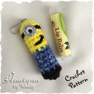 May include: Crocheted Minion lip balm holder with a black eye patch, blue overalls, and a yellow body. The holder is attached to a silver key chain. The lip balm holder is next to a tube of lip balm labeled "All Natural Lip Balm".