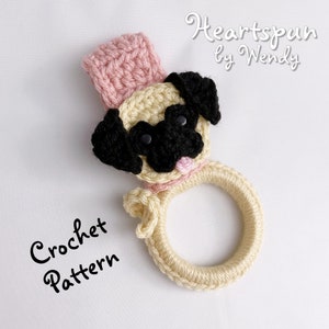 May include: Crochet pattern for a beige and pink pug-shaped baby rattle with black eyes. The text "Crochet Pattern" is written in black on a white background.