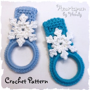 Peut inclure: Deux anneaux de serviette en crochet avec des flocons de neige en embellissement. Les anneaux sont bleus et turquoise, et les flocons de neige sont blancs. Le texte "Heartspun by Wendy" et "Crochet Pattern" est visible sur l'image.