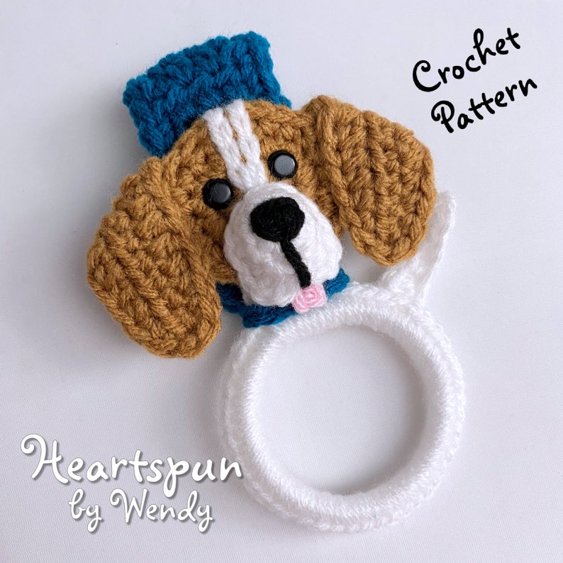 Puede incluir: Porta anillos de crochet con forma de cabeza de perro con un sombrero azul, pelaje marr&oacute;n y blanco y un anillo blanco. El texto "Heartspun by Wendy" est&aacute; debajo del porta anillos. El texto "Crochet Pattern" est&aacute; en la esquina superior derecha de la imagen.