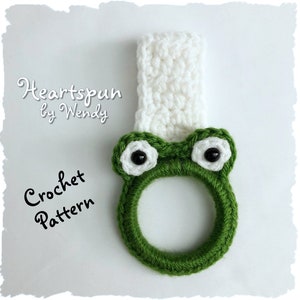May include: Green crocheted frog towel ring with white eyes and a white crocheted top. The text "Heartspun by Wendy Crochet Pattern" is visible in the image.