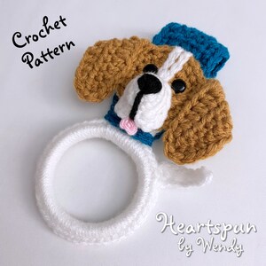 Puede incluir: Porta anillos de cabeza de perro tejido a crochet con un sombrero de marinero azul y blanco. El perro tiene pelo marr&oacute;n, una cara blanca y ojos negros. El texto "Heartspun by Wendy" est&aacute; escrito debajo del perro.