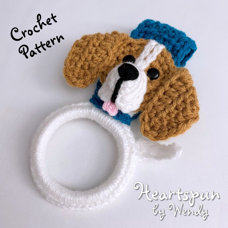 Puede incluir: Porta anillos de cabeza de perro tejido a crochet con un sombrero de marinero azul y blanco. El perro tiene pelo marr&oacute;n, una cara blanca y ojos negros. El texto "Heartspun by Wendy" est&aacute; escrito debajo del perro.