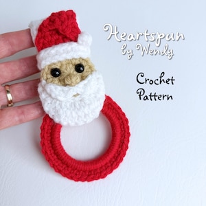May include: A crocheted Santa Claus-themed towel ring. The Santa face is beige with a white beard and a red hat with white trim. The ring is red, and the text "Heartspun by Wendy Crochet Pattern" is visible.