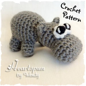 K&ouml;nnte beinhalten: Ein graues, geh&auml;keltes Hippopotamus-Amigurumi-Spielzeug mit schwarzen Augen und wei&szlig;en Akzenten. Das Spielzeug liegt auf einer wei&szlig;en Oberfl&auml;che. H&auml;kelanleitung.