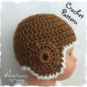 May include: A brown crocheted baby hat with a white crocheted brim. The hat has a circular design on the side. The hat is being worn by a baby.