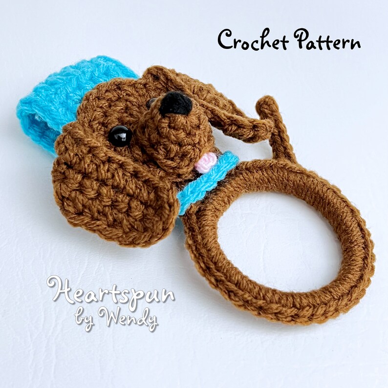 May include: Crocheted brown dog ring holder with a blue and pink bow. The text "Heartspun by Wendy" is below the dog.
