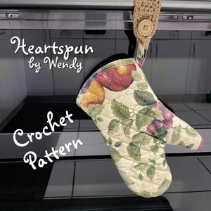 May include: A crocheted oven mitt with a floral pattern featuring apples and grapes. The mitt is hanging from a brown crocheted loop with a wooden button. The text "Heartspun by Wendy Crochet Pattern" is written in white on a black background.