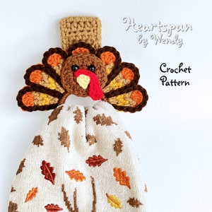 May include: Crochet pattern for a Thanksgiving turkey towel holder. The turkey is brown, orange, and yellow with a red wattle. The towel holder is hanging on a white wall.