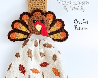 May include: Crochet pattern for a Thanksgiving turkey towel holder. The turkey is brown, orange, and yellow with a red wattle. The towel holder is hanging on a white wall.