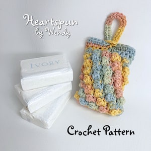 May include: A crochet soap saver with a handle, made in pastel shades of blue, yellow, and pink. The soap saver is designed to hold a bar of soap, such as the Ivory soap bar shown in the image. The text "Heartspun by Wendy" and "Crochet Pattern" are also visible in the image.