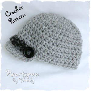 Op de afbeelding: Grijze gehaakte muts met een zwarte knoop en een zwarte en grijze rand. De muts is gelabeld met "Crochet Pattern" en "Heartspun by Wendy".