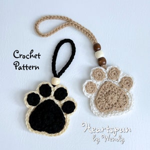 Peut inclure: Deux ornements en forme de patte de chat crochetés, l'un en brun et blanc sur une ficelle blanche, l'autre en noir et blanc sur une ficelle noire. Le texte "Crochet Pattern" et "Heartpsan by Wendy" est visible sur l'image.