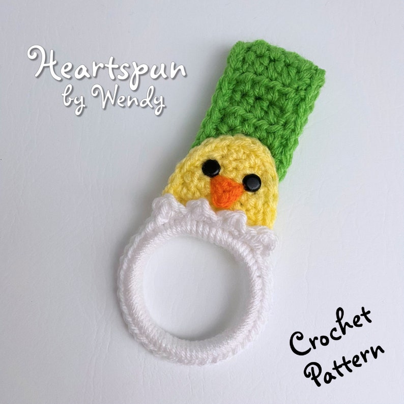 K&ouml;nnte beinhalten: H&auml;kelanleitung f&uuml;r ein gelbes K&uuml;ken mit gr&uuml;nem Hut, das einen wei&szlig;en Ring h&auml;lt. Das Muster hei&szlig;t "Heartspun by Wendy".