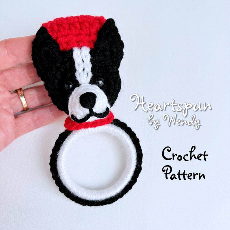 May include: Crochet pattern for a black and white dog with a red hat and scarf. The dog is holding a ring in its mouth. The pattern is called "Heartspun by Wendy".