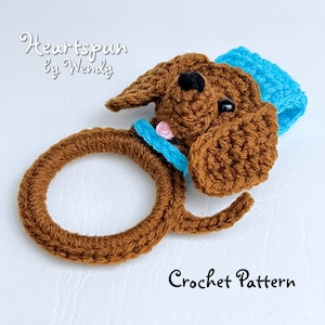 May include: Crochet pattern for a brown dog with a blue hat and a pink flower. The dog is crocheted in the shape of a ring.