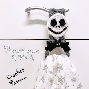 Może przedstawiać: Wzór na szydełku na pierścień na ręcznik z Jackiem Skellingtonem. Pierścień jest szary z czarną kokardką i białą twarzą Jacka Skellingtona. Wzór nazywa się "Heartspun by Wendy".