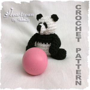 Puede incluir: Un oso panda de crochet sentado junto a un bálsamo labial EOS rosa. El panda es blanco y negro con la nariz y los ojos negros. El bálsamo labial tiene la palabra "eos" impresa.