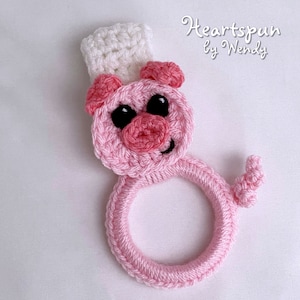 Op de afbeelding: Roze gehaakte varkensringhouder met een witte gehaakte koksmuts.  "Heartspun by Wendy" is in cursief geschreven op een witte achtergrond.