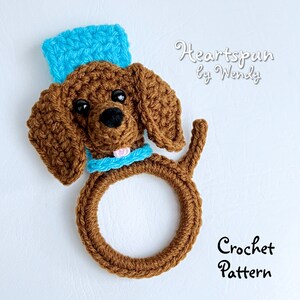May include: Crochet pattern for a brown dachshund dog wearing a blue hat and a blue collar. The dog is holding a brown ring in its mouth. The pattern is called "Heartspun by Wendy".