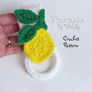 May include: A crocheted lemon-shaped dishcloth with green leaves. The dishcloth is white with yellow and green crocheted details. The text "Heartspun by Wendy Crochet Pattern" is visible in the background.