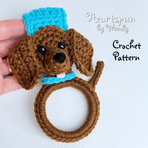 May include: Crochet pattern for a brown dachshund dog with a blue hat and collar. The dog is holding a ring in its mouth. The pattern is called "Heartspun by Wendy".