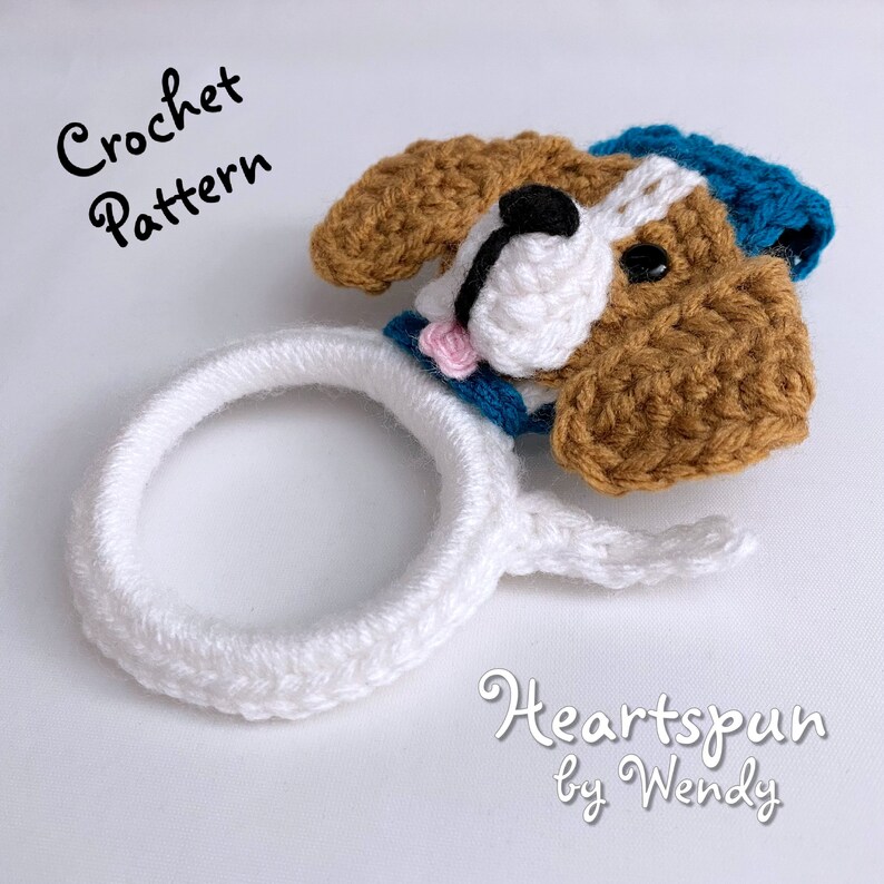 Puede incluir: Patr&oacute;n de crochet para un perro blanco y marr&oacute;n con un sombrero azul. El perro tiene una flor rosa en la boca. El patr&oacute;n se llama "Heartspun by Wendy".
