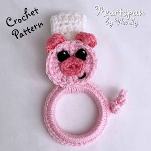May include: A pink crocheted pig with a white crocheted hat. The pig has black eyes and a pink snout. The pig is holding a pink crocheted ring.