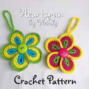 Puede incluir: Dos adornos de flores de ganchillo con un lazo para colgar. Una flor es amarilla, verde y azul, y la otra es amarilla, verde y rosa. Las flores tienen un botón en el centro. El texto "Heartspun by Wendy Crochet Pattern" está debajo de las flores.