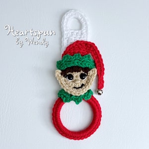Puede incluir: Un adorno de elfo de ganchillo con un gorro rojo, un gorro verde y una cara marrón. El elfo sostiene un anillo rojo con un lazo blanco en la parte superior. El adorno tiene una campana plateada unida al gorro.
