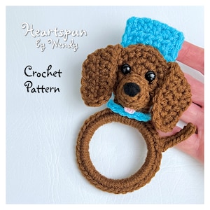 Puede incluir: Juguete de perro salchicha marrón tejido a crochet con un sombrero azul y una pajarita azul. El juguete tiene forma de anillo y tiene una cola que forma parte del anillo. El texto "Heartspun by Wendy Crochet Pattern" es visible en la imagen.