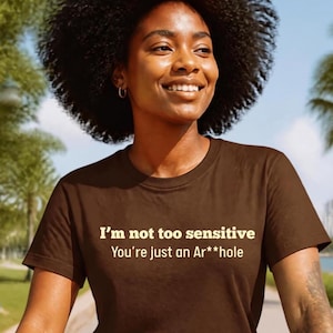 May include: A dark brown short-sleeved t-shirt featuring the text in light yellow lettering that reads: I'm not too sensitive You're just an Ar**hole. The shirt has a classic crew neckline and a standard fit, providing a simple and direct design.