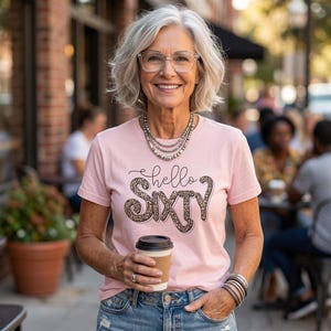 May include: A light pink short-sleeved t-shirt featuring the text "hello SIXTY" in a black script and leopard print block font. The shirt is paired with blue denim jeans, a multi-strand silver beaded necklace, and a stack of silver bracelets.