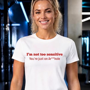 May include: A white short-sleeved t-shirt featuring red text printed on the chest that reads, I'm not too sensitive You're just an Ar**hole. The garment has a classic crew neckline and a straight hem, providing a simple and clean design.